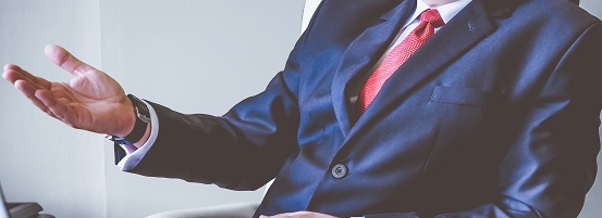 Mann in dunkelblauem Anzug, weißem Hemd und roter Krawatte diskutiert mit erhobener Hand.