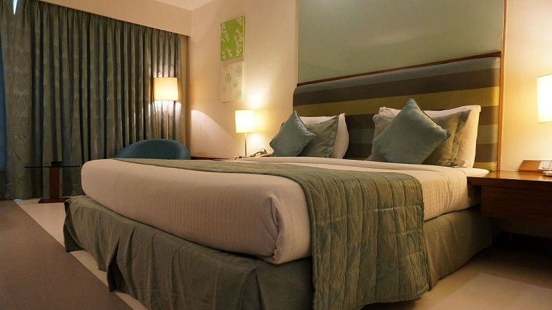 Hotelzimmer mit Bett, zugezogenen Vorhängen, zwei Gemälden an der Wand, jeweils drei Kissen auf jeder Seite der Matratze und drei Stehlampen.