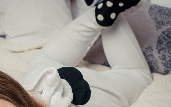 Junge Frau liegt in einem weißen Schlafanzug und mit schwarzen Socken mit weißen Punkten auf dem Bett.