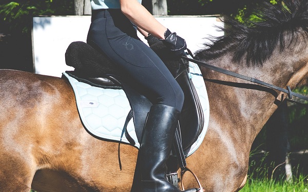 Reiterin in Reitermontur sitzt mit schwarzen Stiefeln, schwarzer Hose, schwarzen Handschuhen und hellem Top auf einem braunen Pferd und reitet.