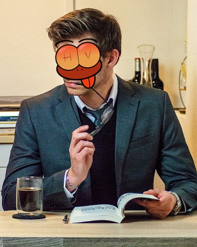 Spanking Treffen: Spanker in einem grauen Anzug mit schwarzem Pullunder, grauer Krawatte und weißem Hemd. Er blickt zur Seite und hat den Flügel seiner Brille im Mund